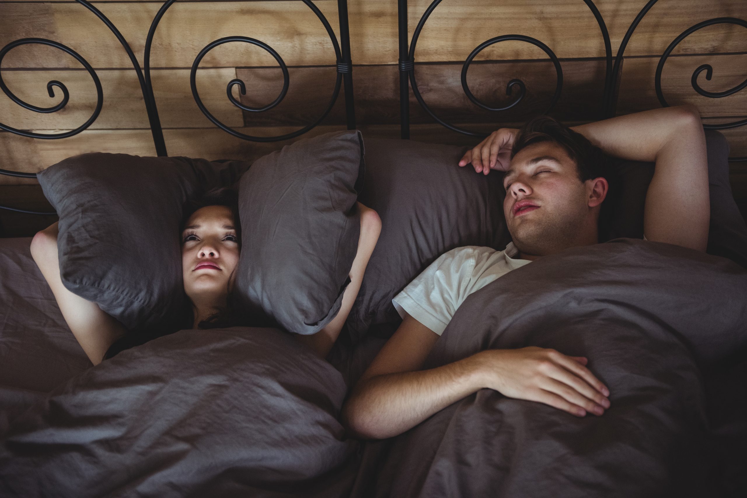Annoyed woman covering her ears with pillows to block out snoring in bedroom at home